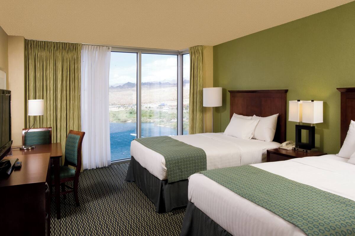 Ein Hotelzimmer mit zwei Betten, einem Schreibtisch und einem Stuhl. Es gibt ein großes Fenster mit Vorhängen, eine Lampe und einen Blick auf eine Landschaft.