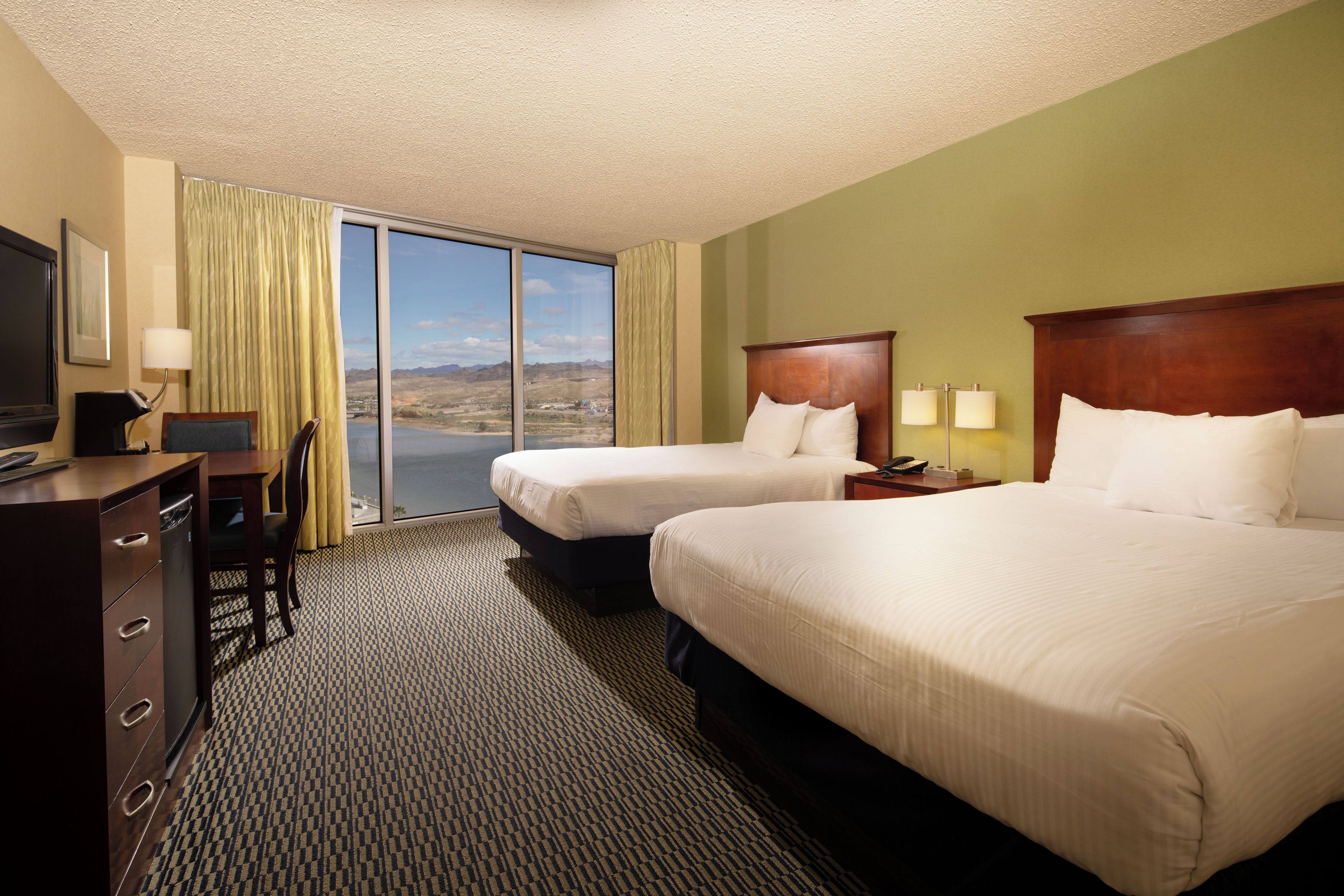 A hotel room with two beds, a desk with a computer, a TV, and a view of a mountainous landscape.