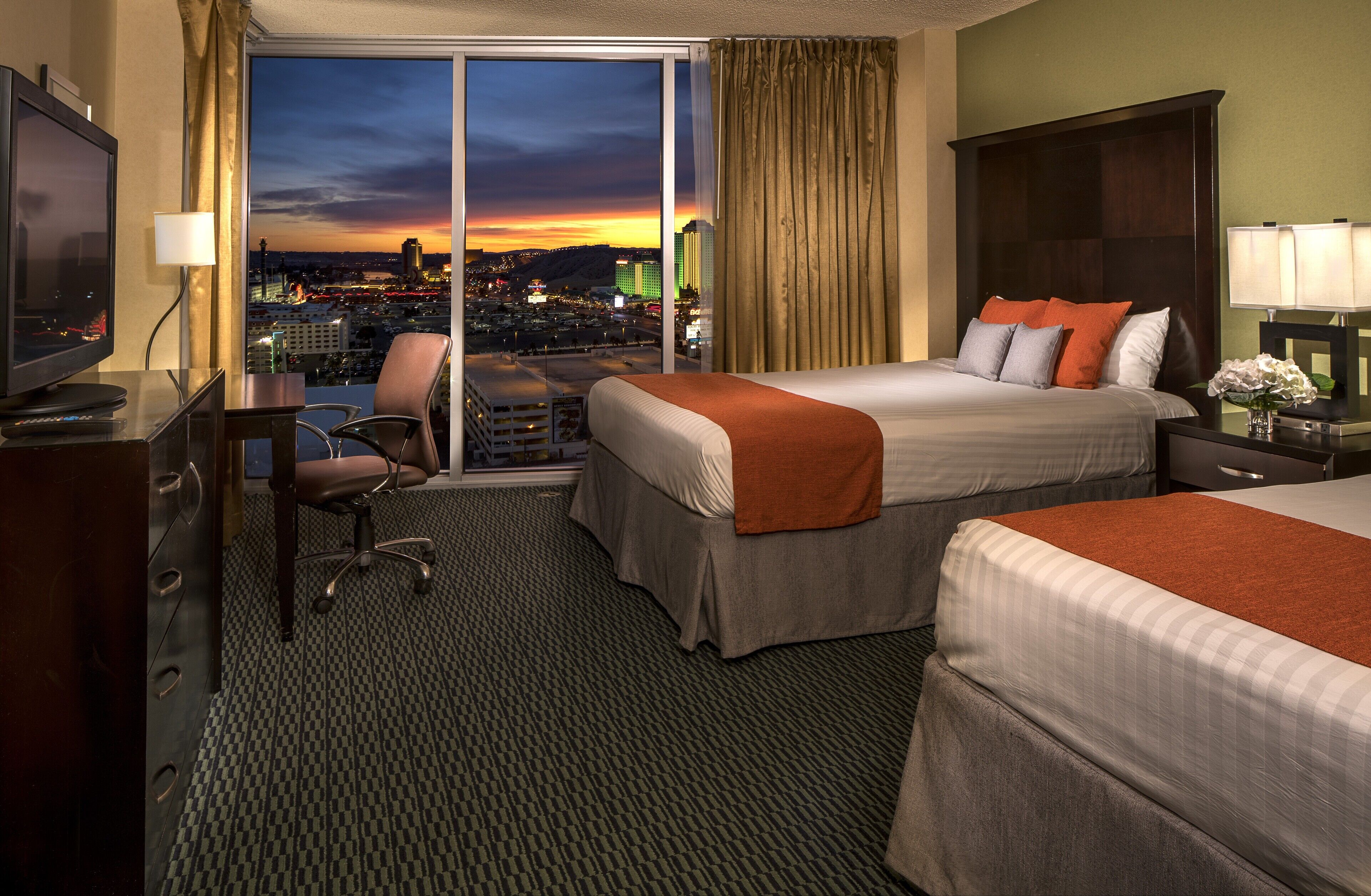 A hotel room with two beds, a desk with a TV, a chair, and a view of a cityscape at sunset.