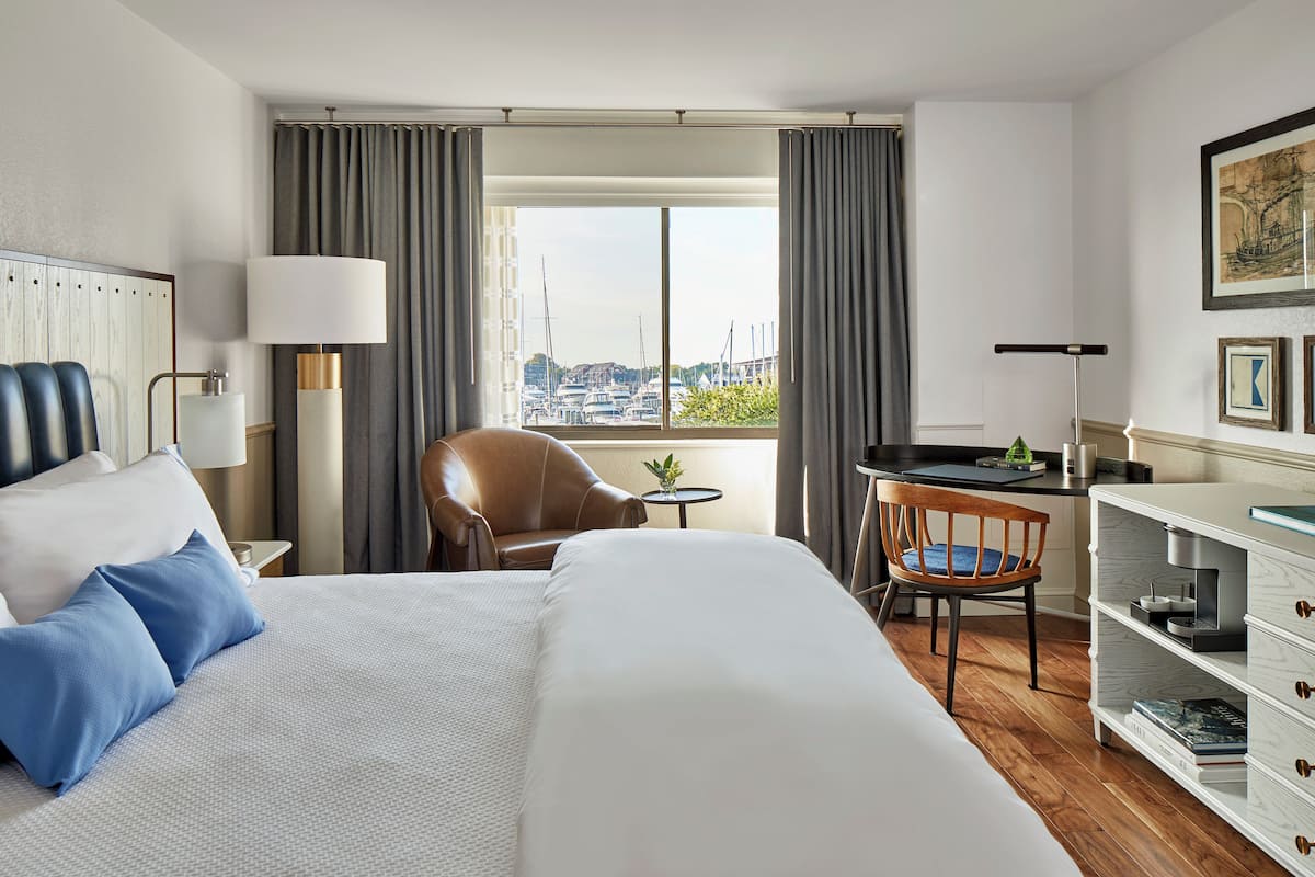 A hotel room with a large bed, a desk, a chair, and a view of boats through the window.