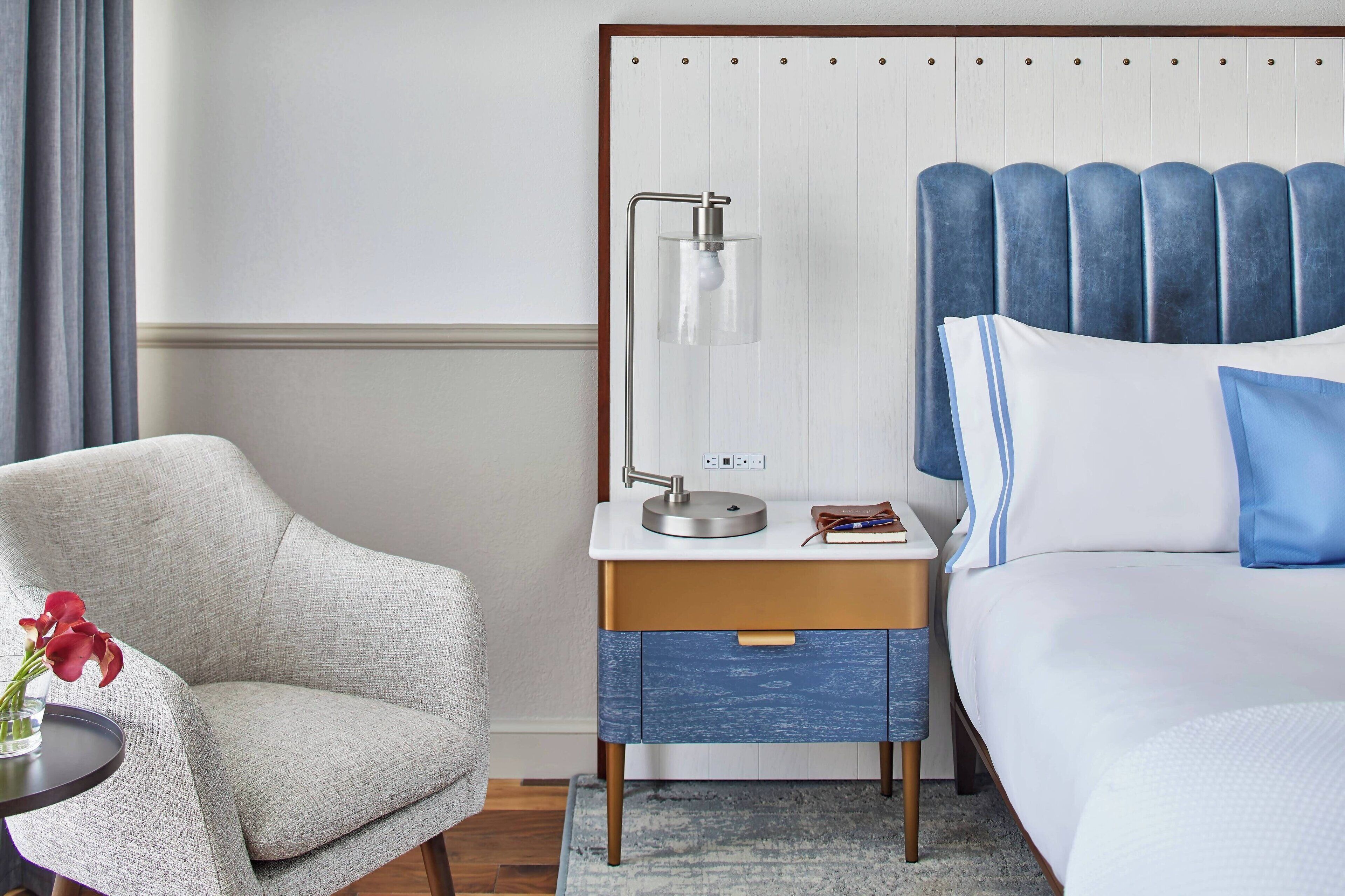 A hotel room with a bed, a nightstand with a lamp, a chair, and a small table with a vase of flowers.