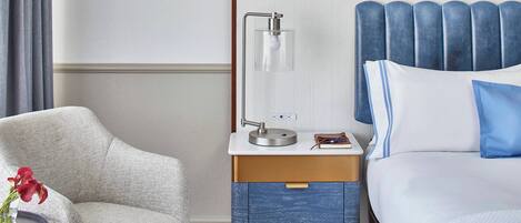 A hotel room with a bed, a nightstand with a lamp, a chair, and a small table with a vase of flowers.