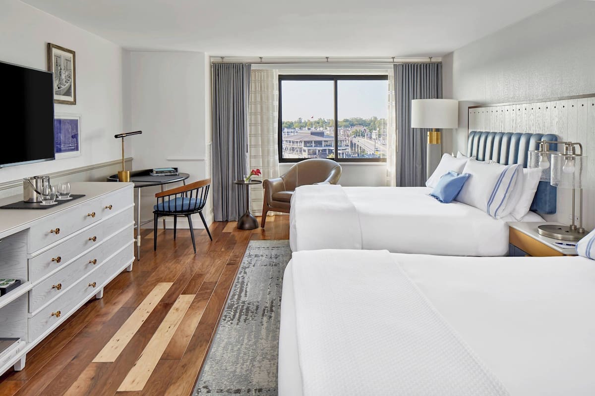 A hotel room with two beds, a desk, a chair, a TV, and a window with a city view.
