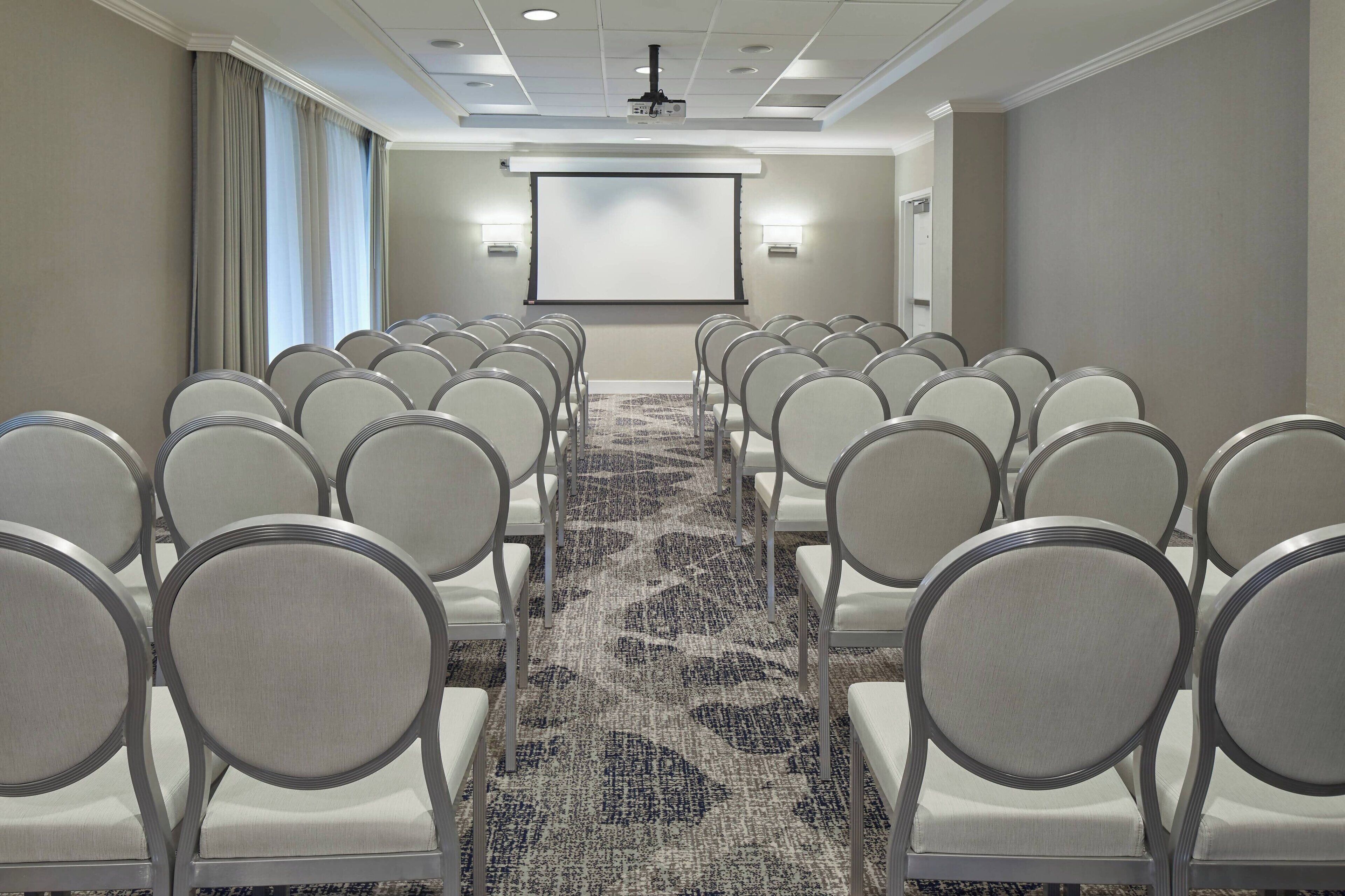 A conference room with rows of chairs, a carpeted floor, a projector screen, and a ceiling-mounted projector.