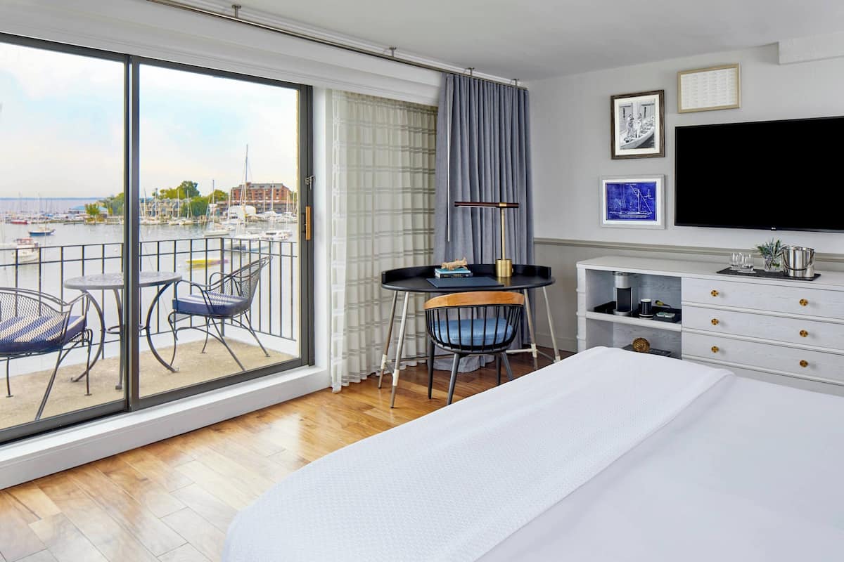 A hotel room with a large bed, a TV, a desk with a chair, and a balcony with a view of the marina.