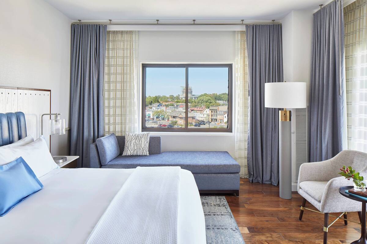 A hotel room with a large bed, a sofa, a chair, a lamp, and a view of the city through a window.