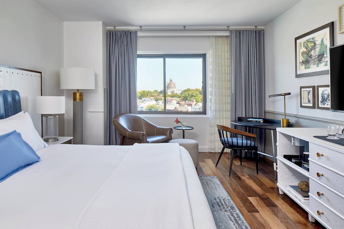 A hotel room with a large bed, a desk, a chair, a TV, and a window with a view of buildings.