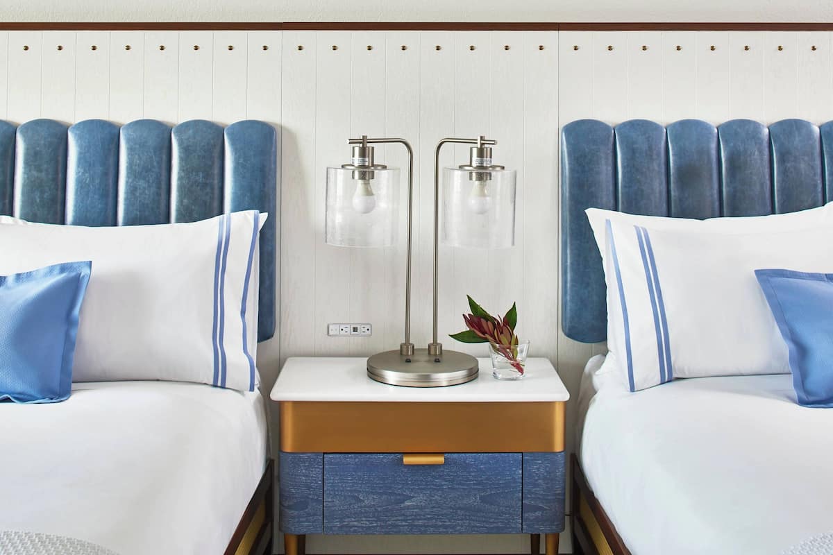 A hotel room with two beds, a nightstand with a lamp, and a small drawer.