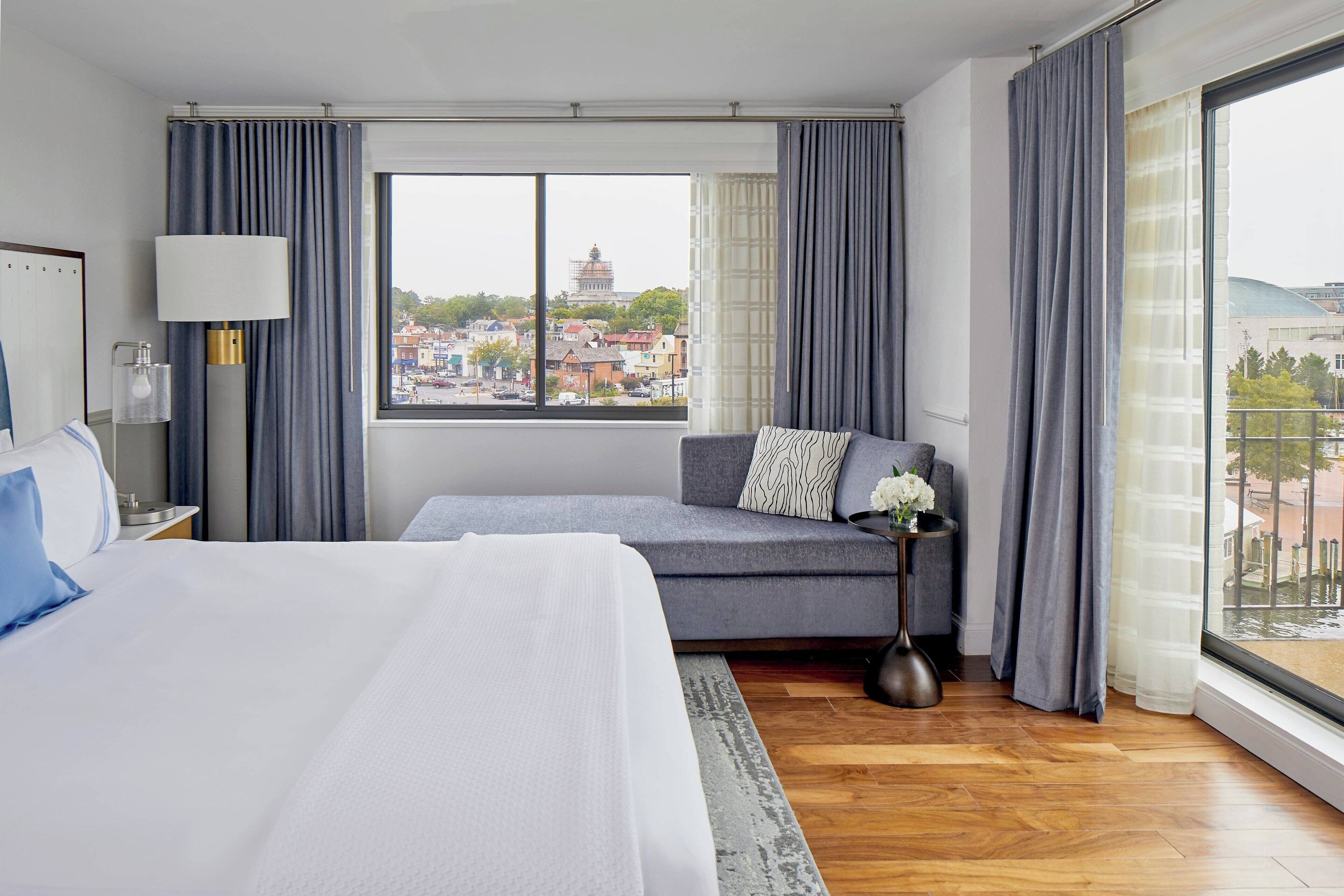 Une chambre d’hôtel moderne avec un grand lit, un canapé et une vue sur le paysage urbain, visible à travers une grande fenêtre.