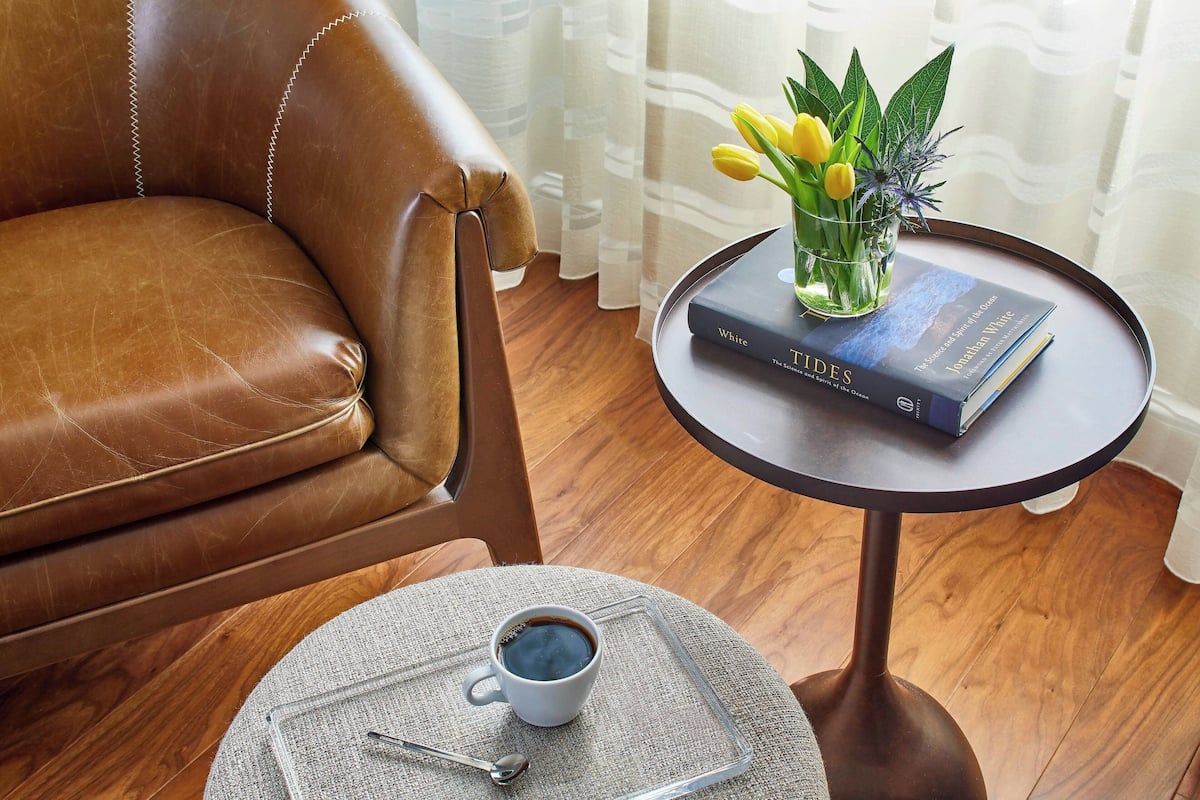 A leather armchair, a small round table with a book and a glass vase with flowers, and a cup of coffee on a coaster.