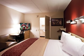 Ett hotellrum med en säng, ett skrivbord med en tv, en stol, en dörr och en väggmonterad lampa.