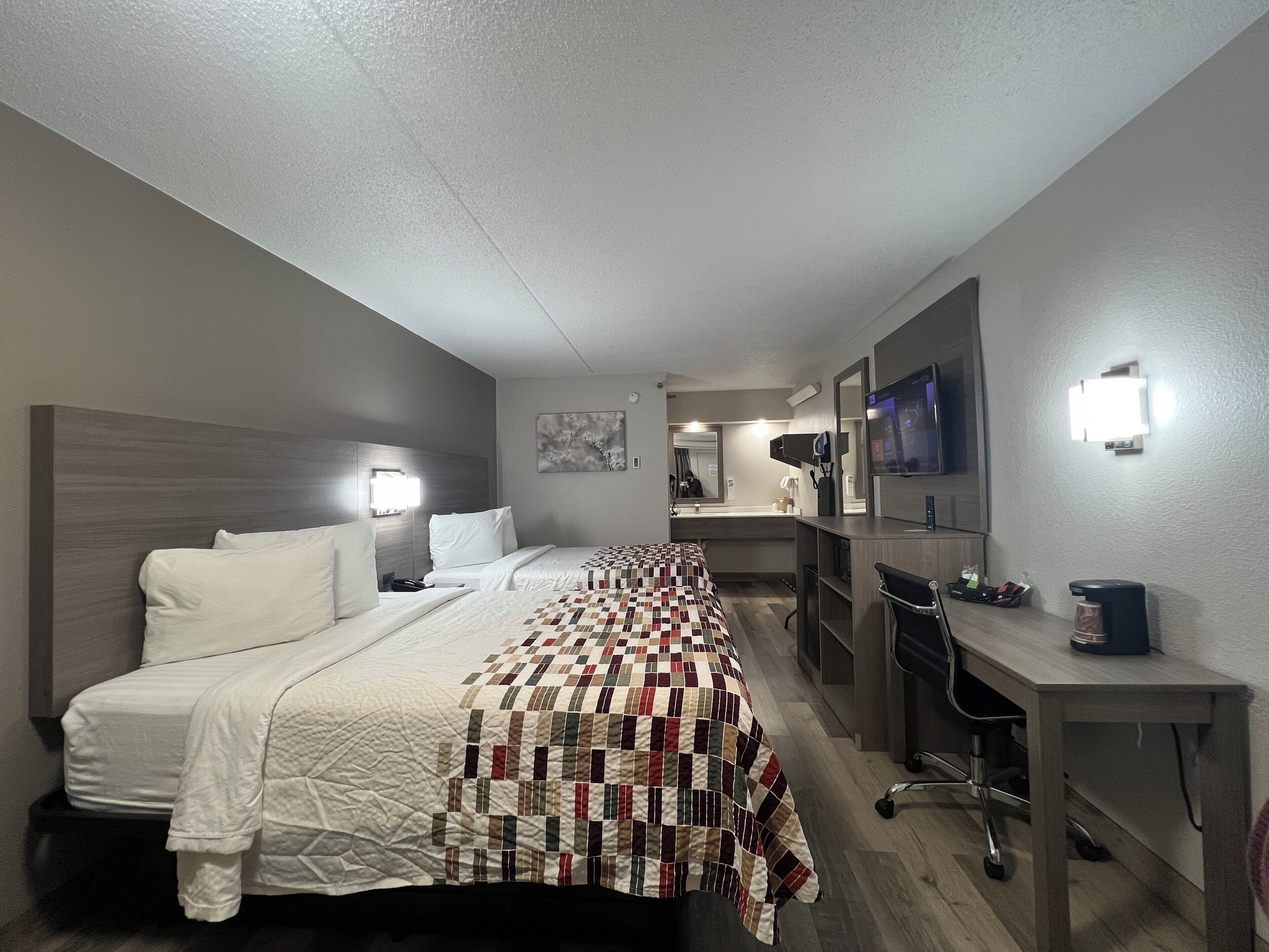 A hotel room with a bed, a desk, a chair, a TV, and a bathroom visible in the background.