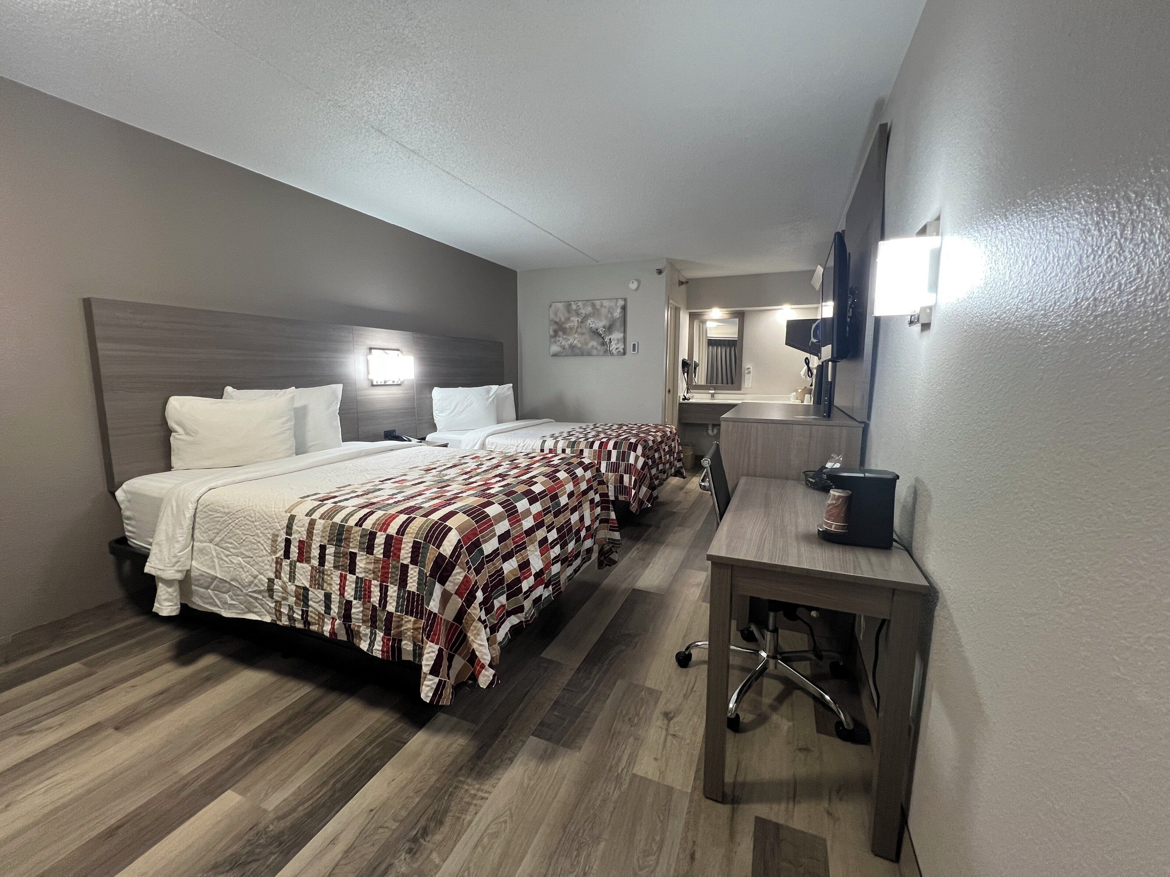A hotel room with two beds, a desk, a mirror, and a small kitchenette.