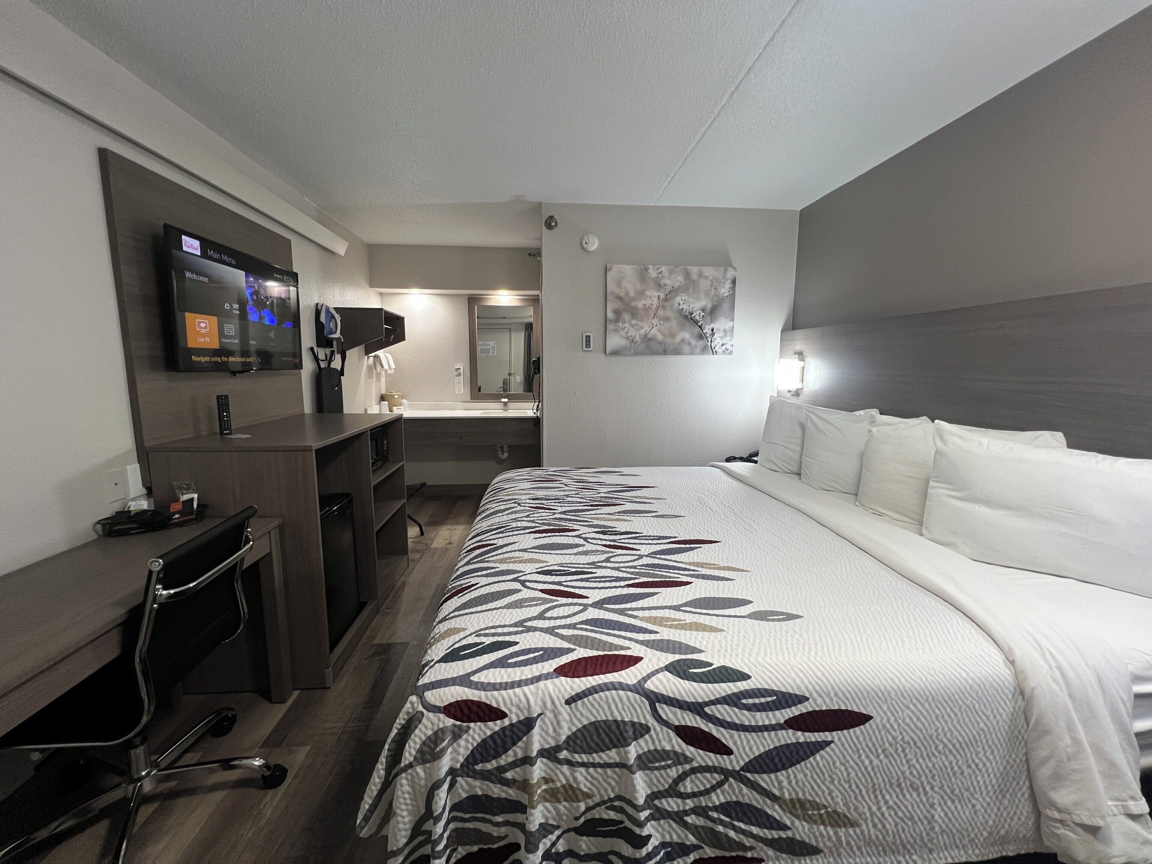 Ett hotellrum med en säng, ett skrivbord, en tv, en spegel och en tavla på väggen.