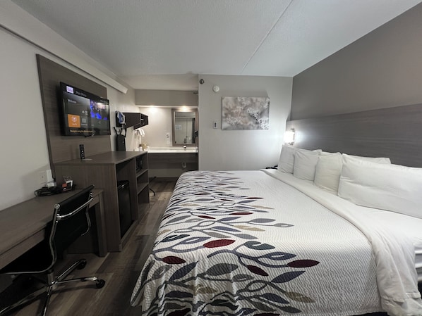 Hotellihuone, jossa on sänky, työpöytä, televisio, peili ja taulu seinällä.