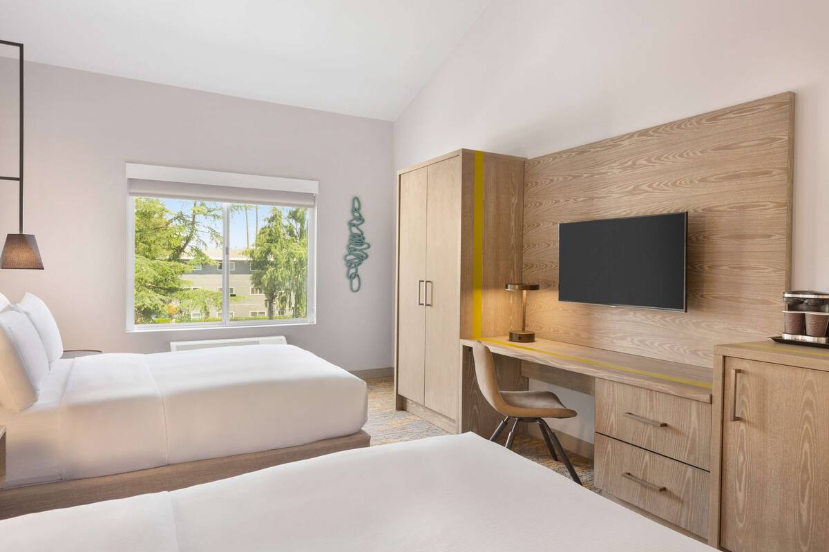 Quarto de hotel moderno com duas camas, uma escrivaninha, uma cadeira, uma televisão e uma janela com vista para o verde.