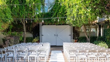Een terras met rijen witte stoelen en een witte tafelkleed, omgeven door groen en een wit hek.