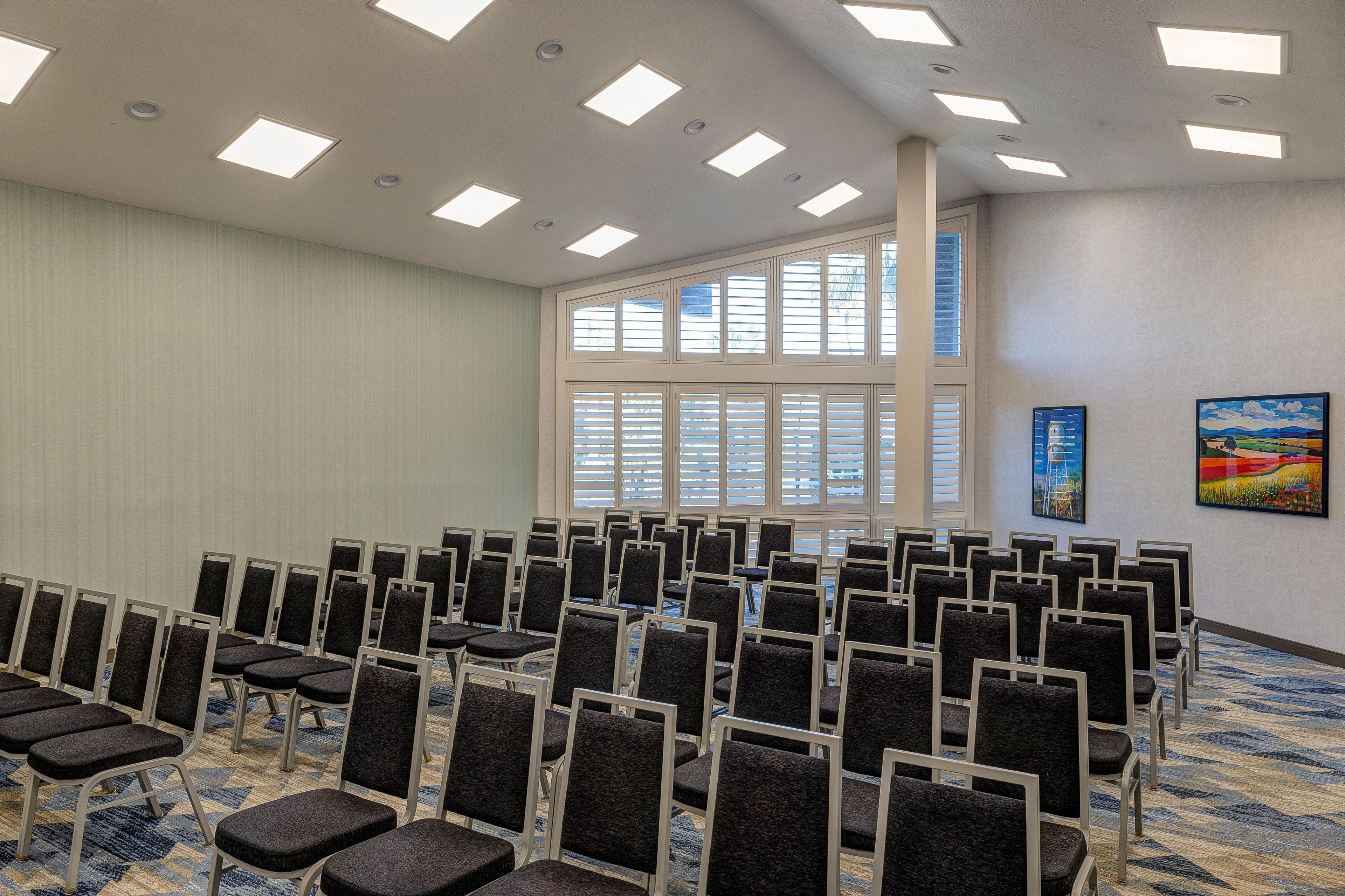 Konferenssihuone, jossa on riveissä tuoleja, suuret ikkunat ja kattovalaisimet.