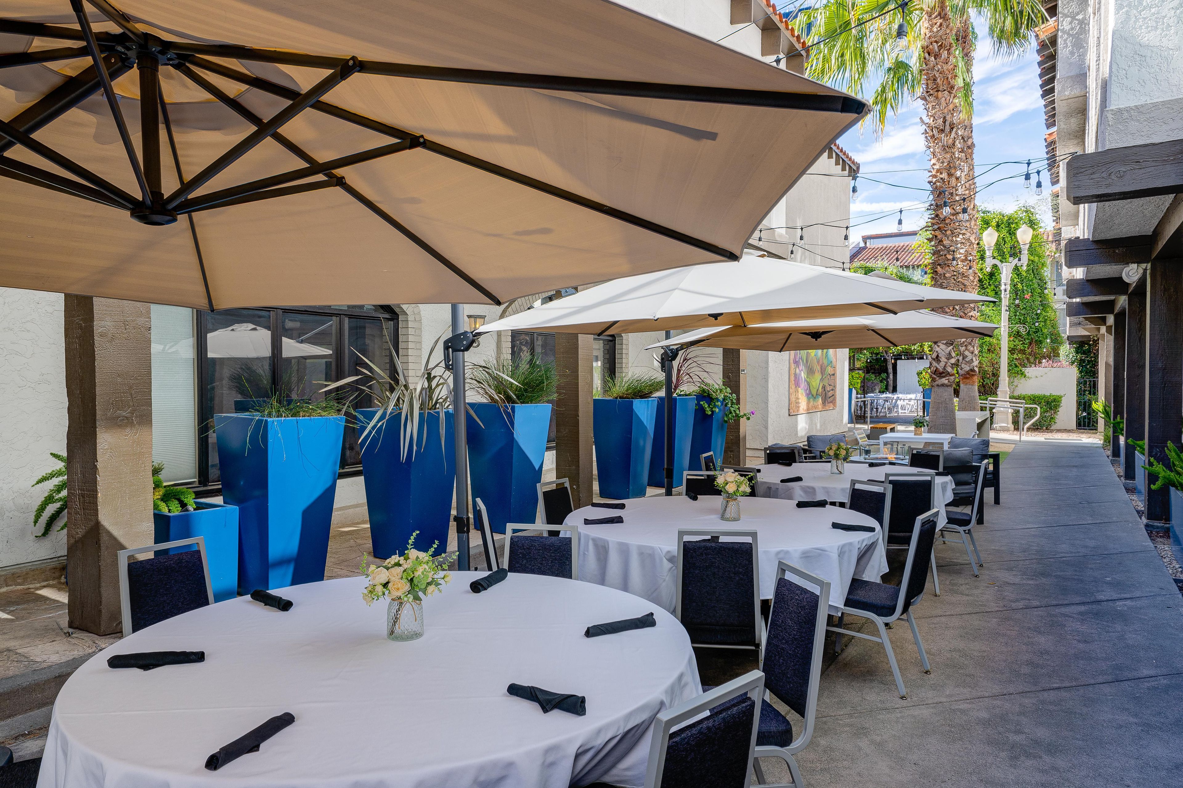 Un espace extérieur aménagé pour les repas, avec des tables et des parasols, entouré de jardinières bleues et bordé d’un chemin.