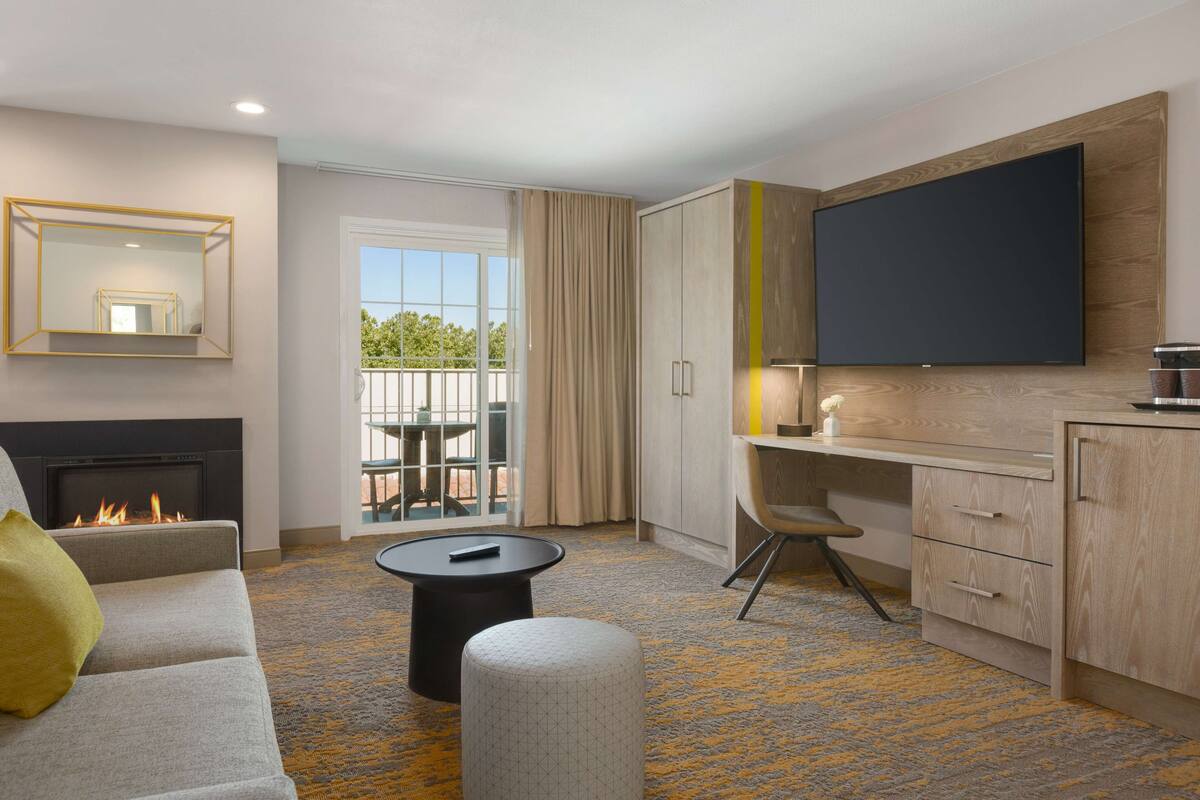 Quarto de hotel moderno com sofá, pequena mesa redonda, lareira, varanda com vista, escrivaninha e TV de tela plana fixada na parede.