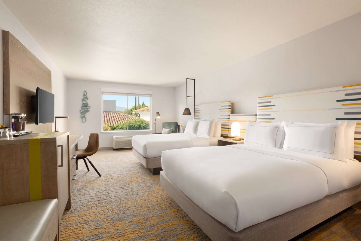 Quarto de hotel com duas camas, uma escrivaninha, uma cadeira, televisão e janela com vista para o exterior.