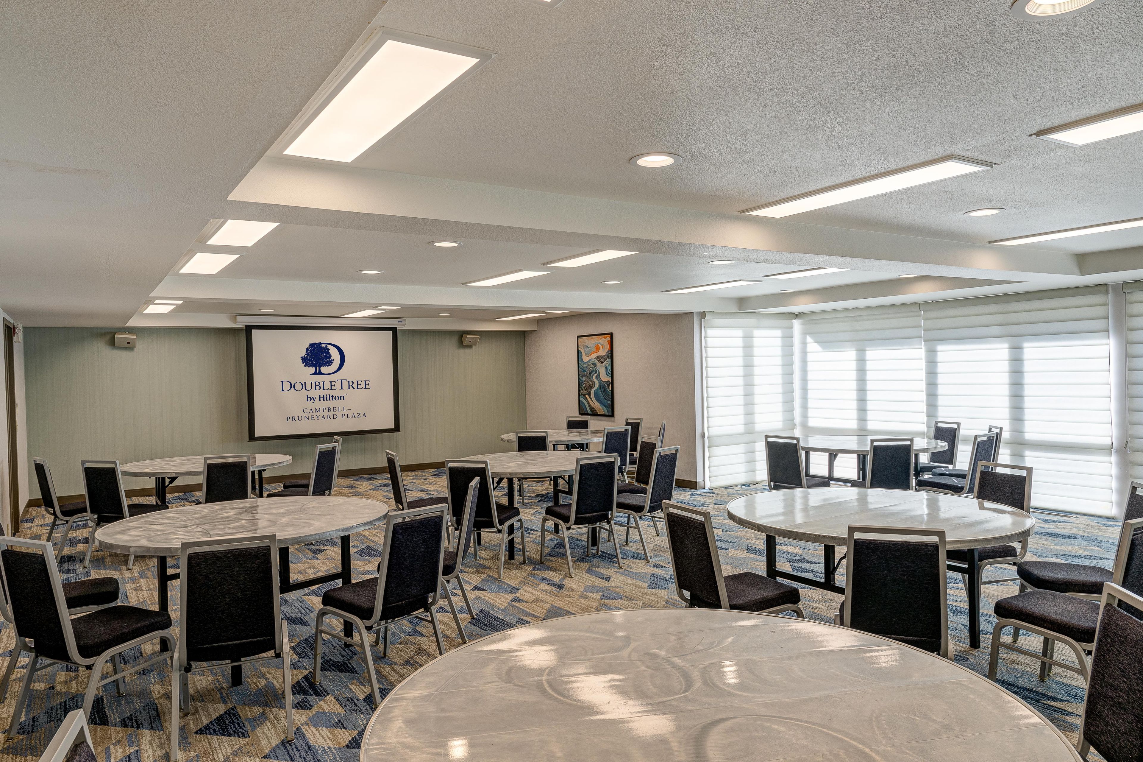 A conference room with multiple round tables and chairs, a projector screen, and a large window with blinds.
