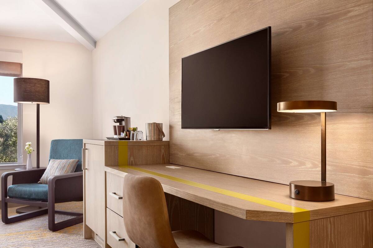 Quarto de hotel moderno com escrivaninha de madeira, televisão de tela plana na parede, poltrona confortável e luminária.