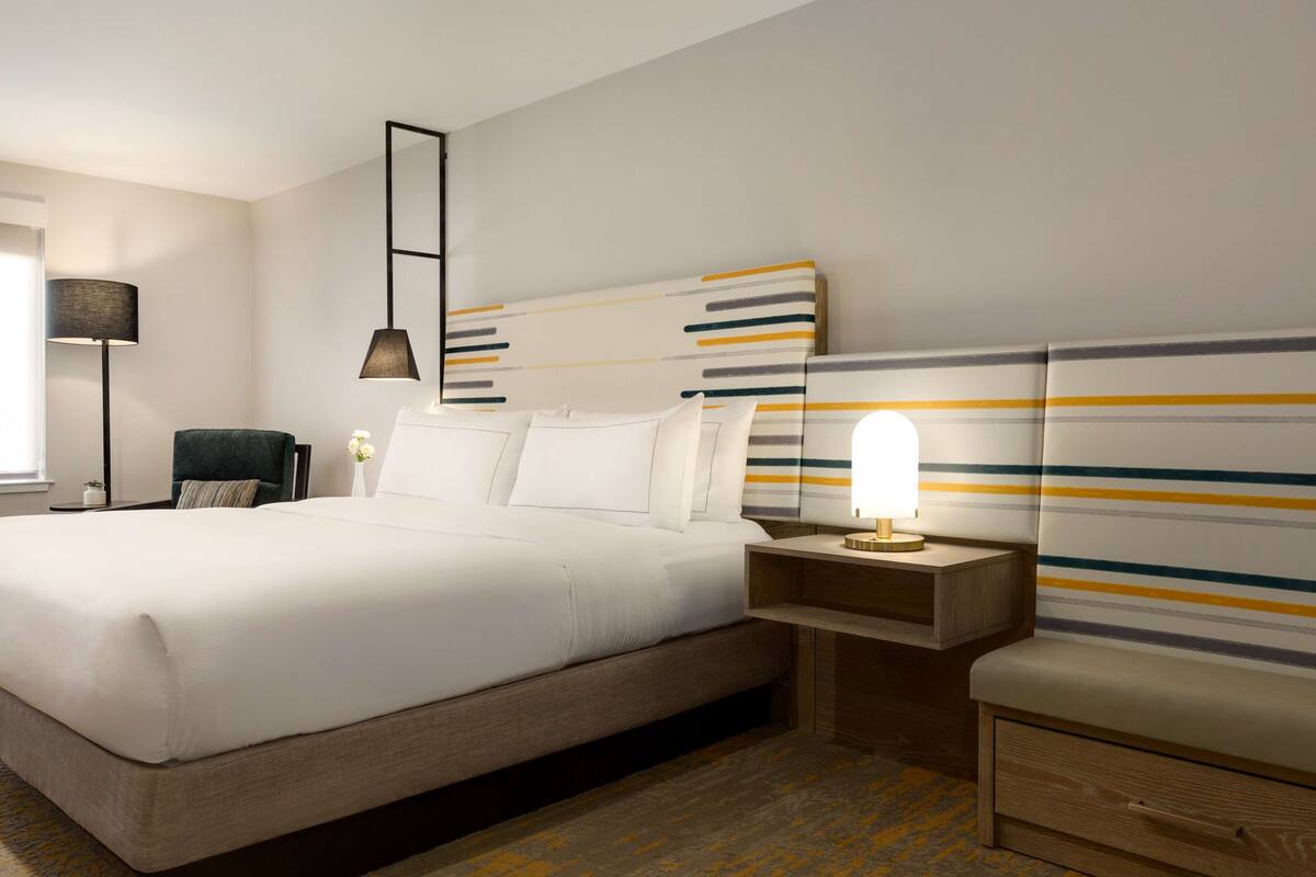 Quarto de hotel com uma cama grande, mesas de cabeceira, uma luminária e uma cadeira.