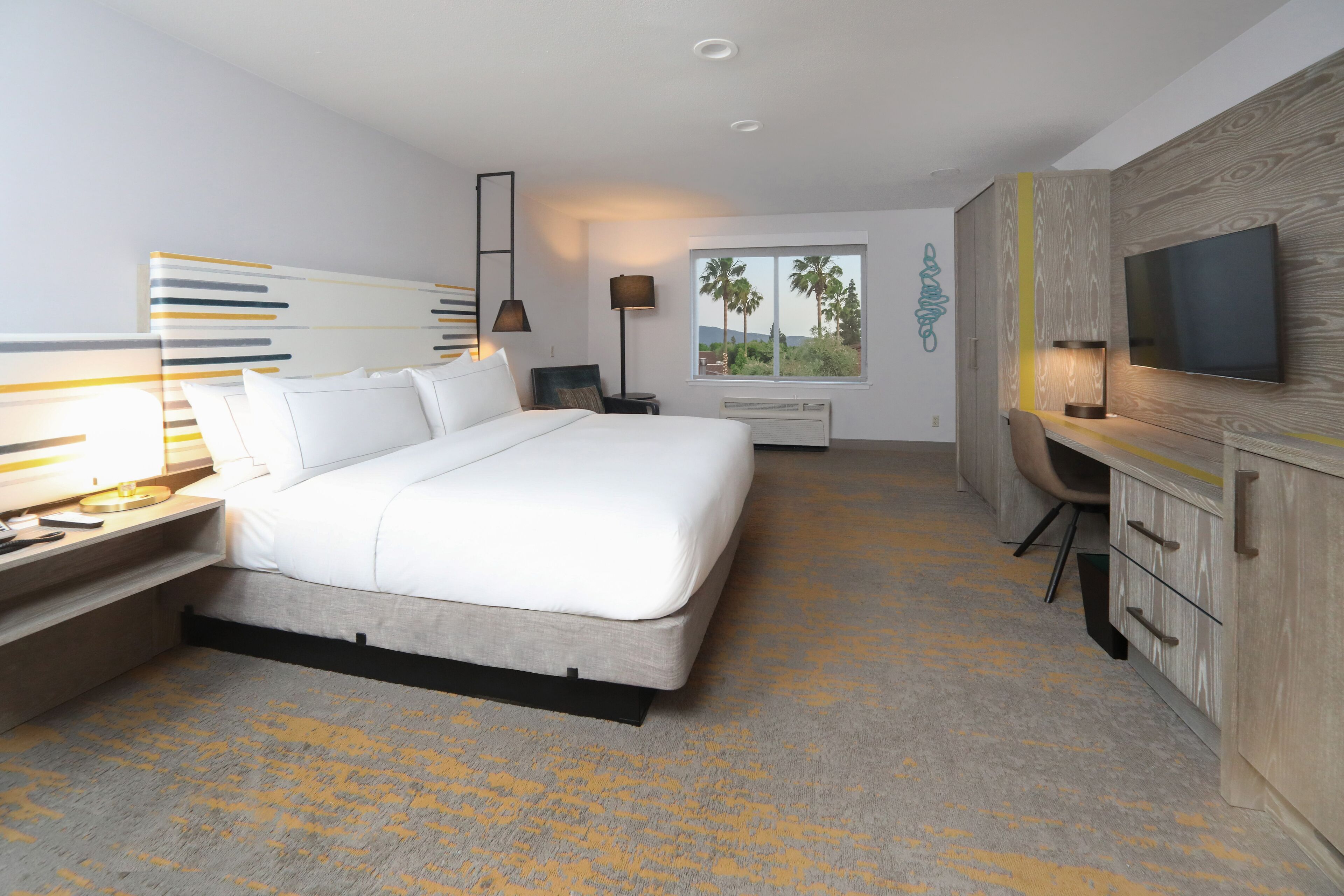 A modern hotel room with a large bed, a desk, a TV, and a window with a view of palm trees.