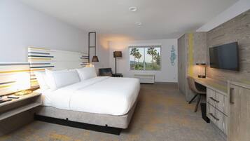 A modern hotel room with a large bed, a desk, a TV, and a window with a view of palm trees.