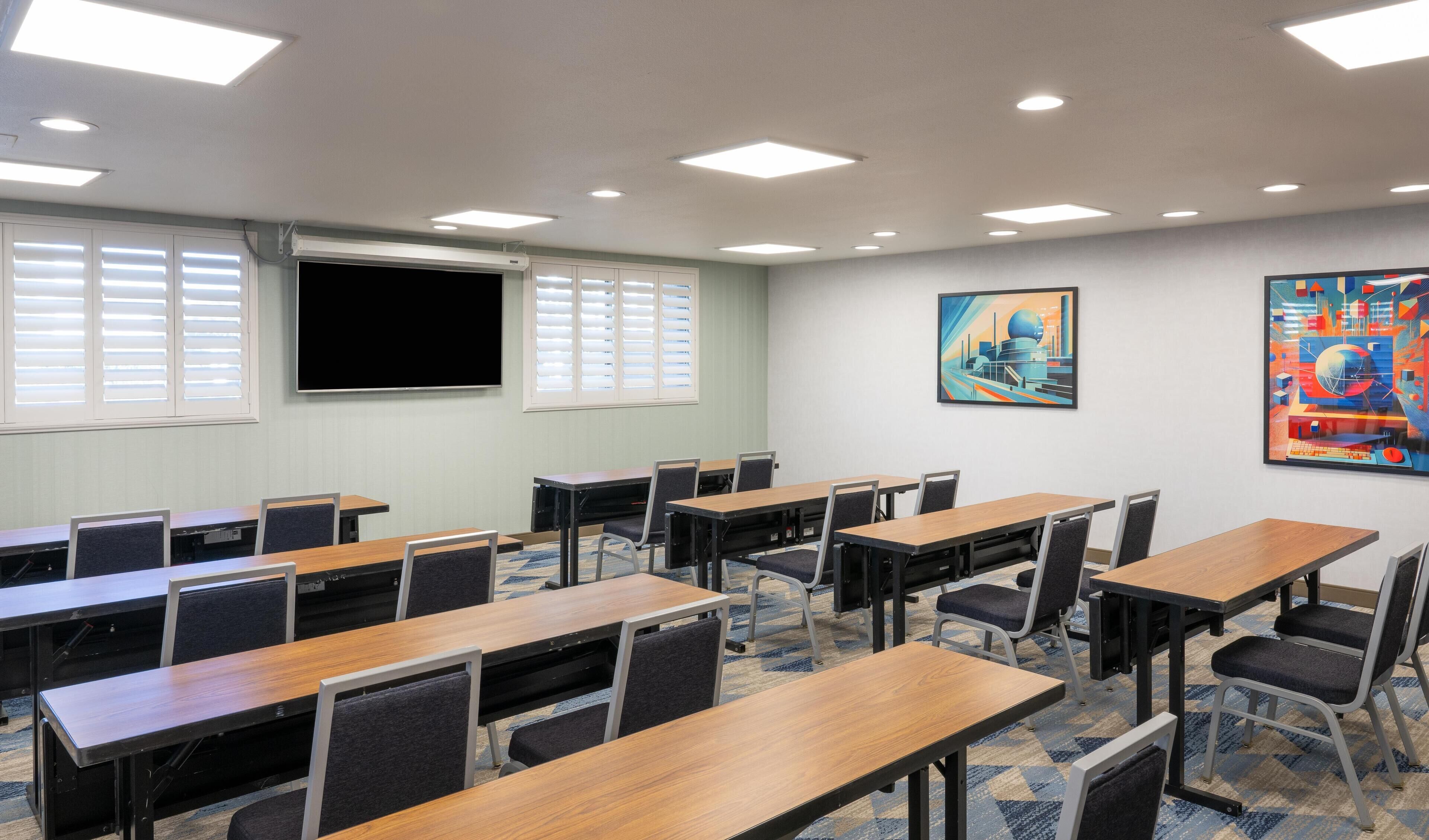 Un salón de conferencias con varias mesas y sillas, una pantalla grande y dos ventanas con persianas.