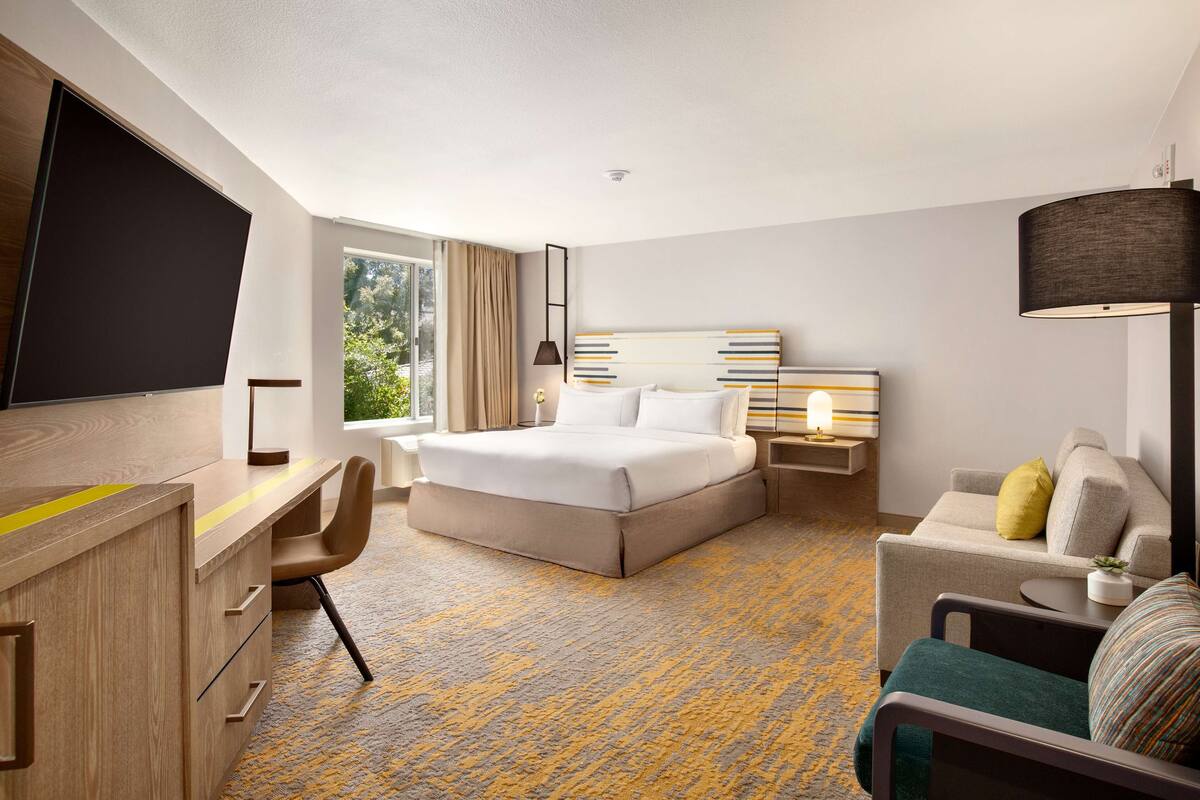 Quarto de hotel com cama, escrivaninha, cadeira, sofá e uma janela grande.