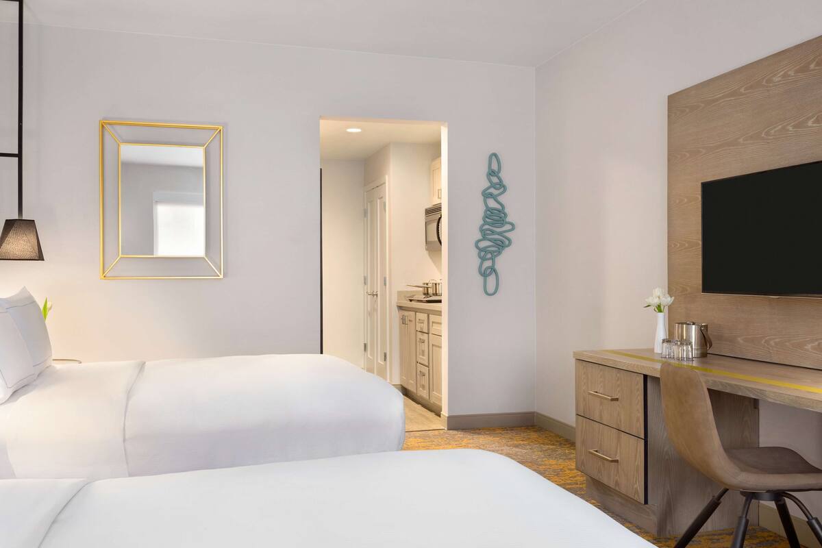 Quarto de hotel com duas camas, uma escrivaninha, televisão e banheiro visível por uma porta aberta.