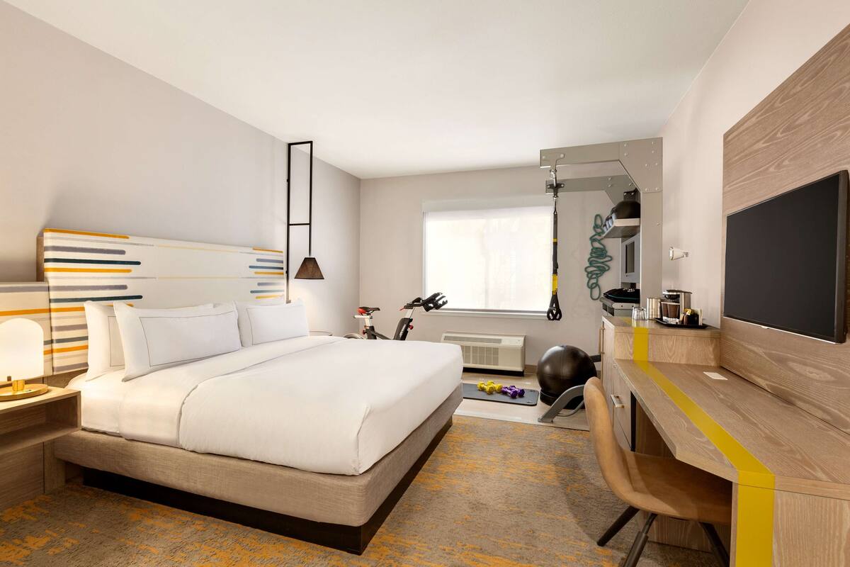 Quarto de hotel moderno com uma cama grande, uma mesa com cadeira, televisão e janela com cortinas.