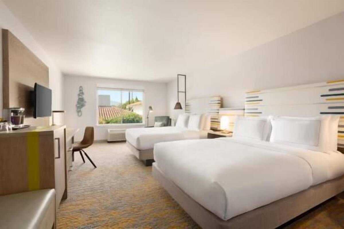 Quarto de hotel com duas camas, uma escrivaninha, uma cadeira, uma televisão e uma janela com vista.