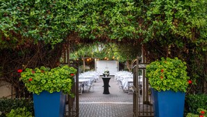 Un jardín al aire libre con zona pavimentada, mesas, sillas y una mesa central adornada con un jarrón de flores.