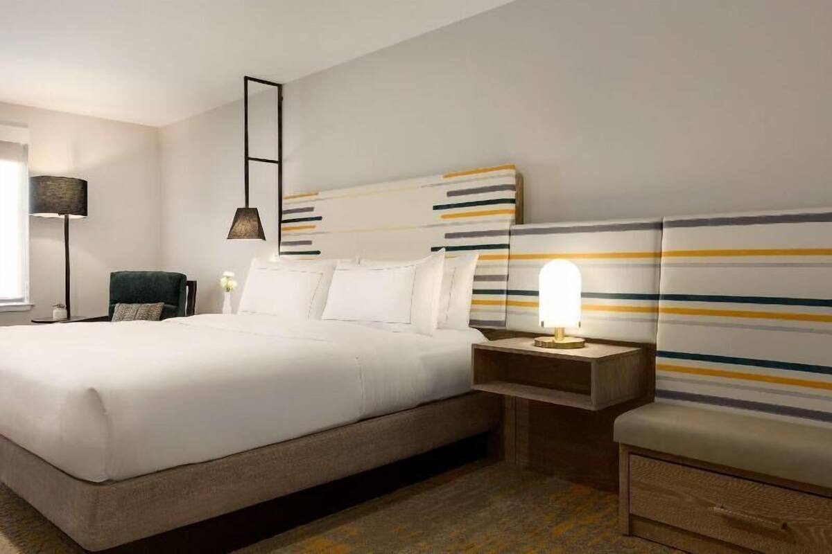 Quarto de hotel com uma cama grande, mesas de cabeceira, uma luminária e uma cadeira.