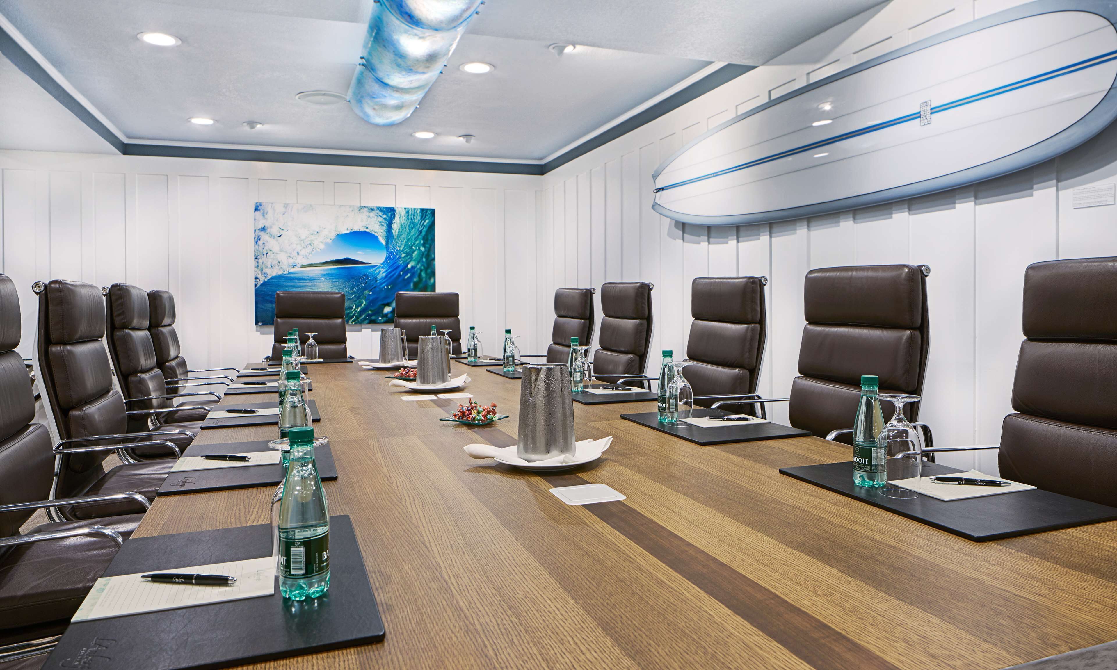 A conference room with a long wooden table, brown leather chairs, water bottles, and a surfboard decoration on the wall.