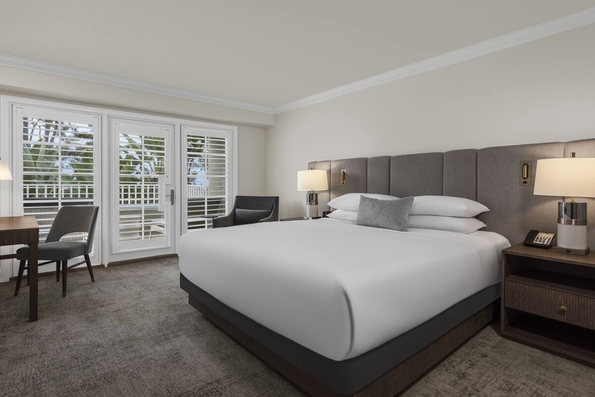 Habitación de hotel con una cama grande, un escritorio con silla, una mesita de noche con lámpara y un ventanal corredero que da a un balcón.
