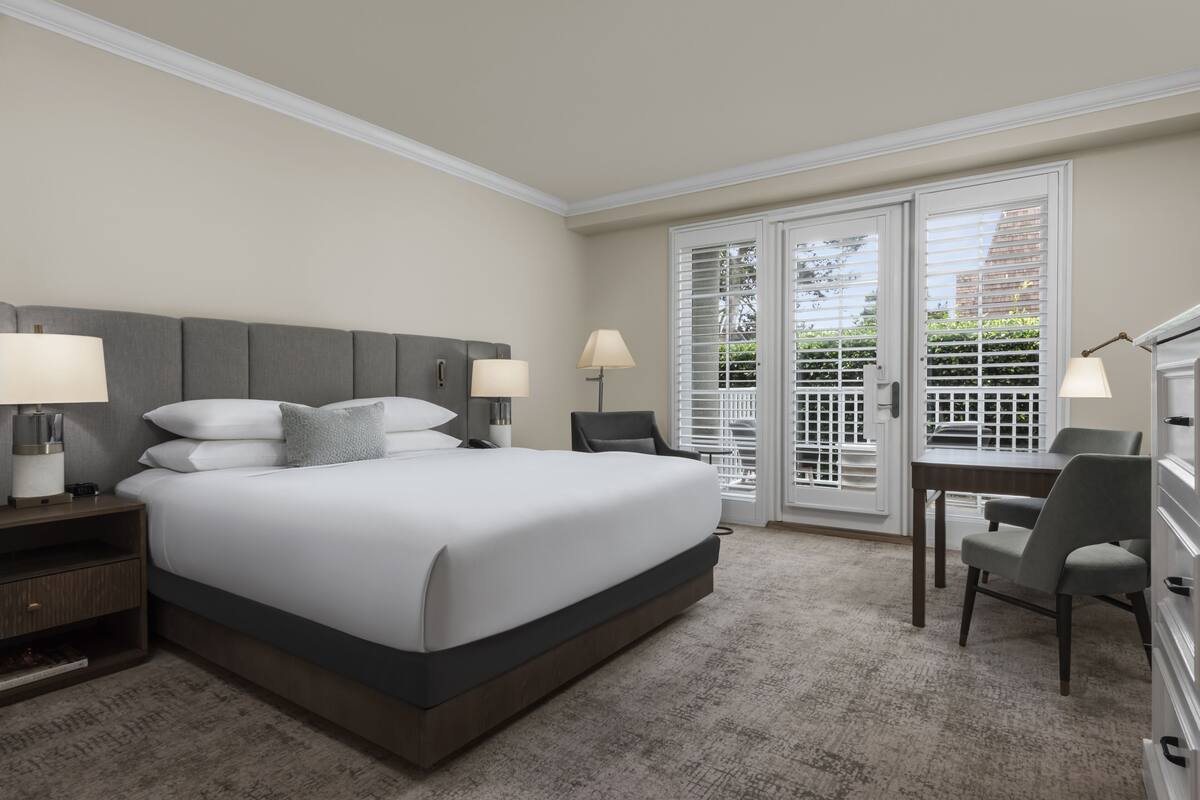 Habitación de hotel con una cama grande, dos mesitas de noche con lámparas, un escritorio con una silla y vistas a través de puertas de cristal.