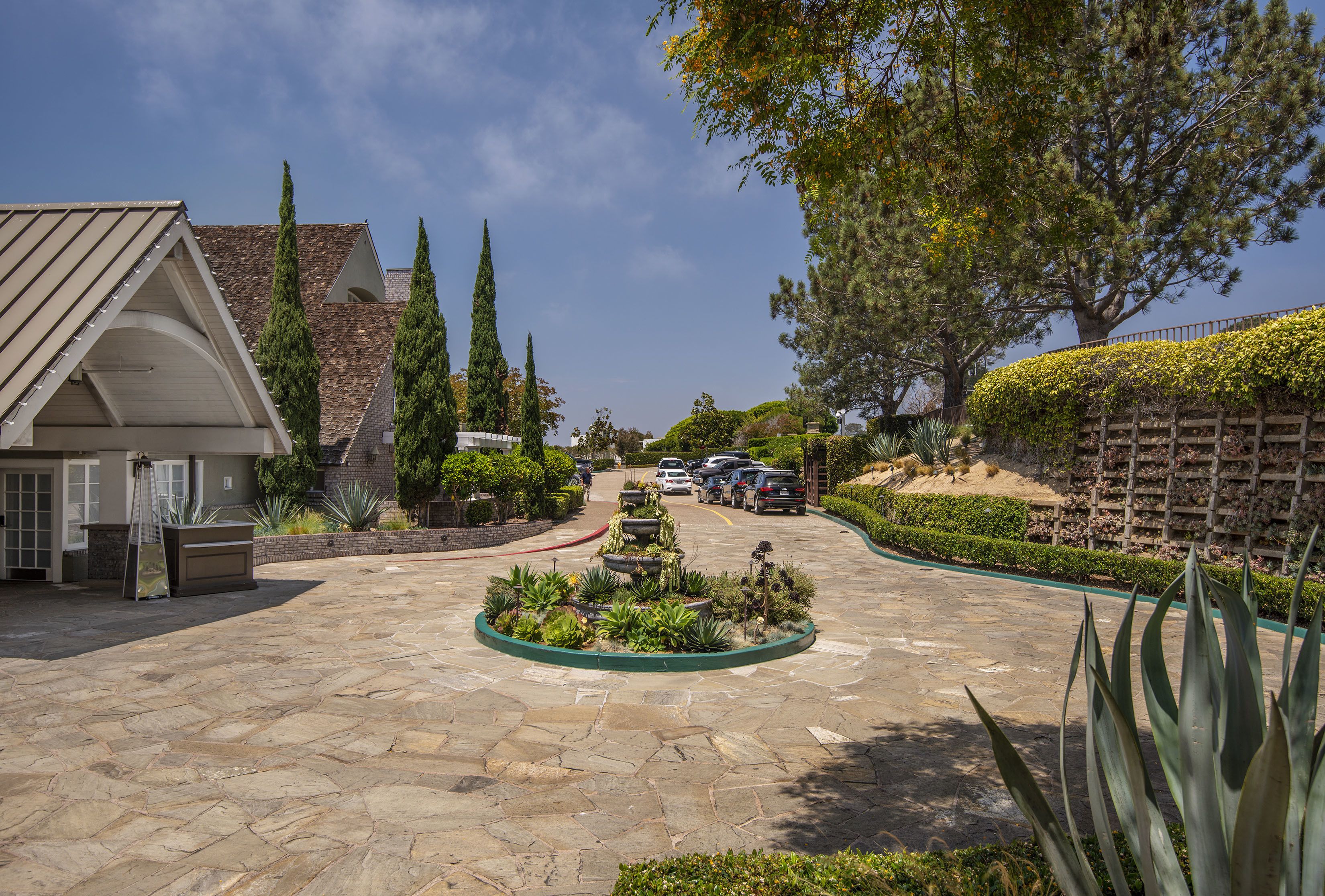 A stone-paved courtyard with a central garden, a building with a sloped roof, and a fence with climbing plants.
