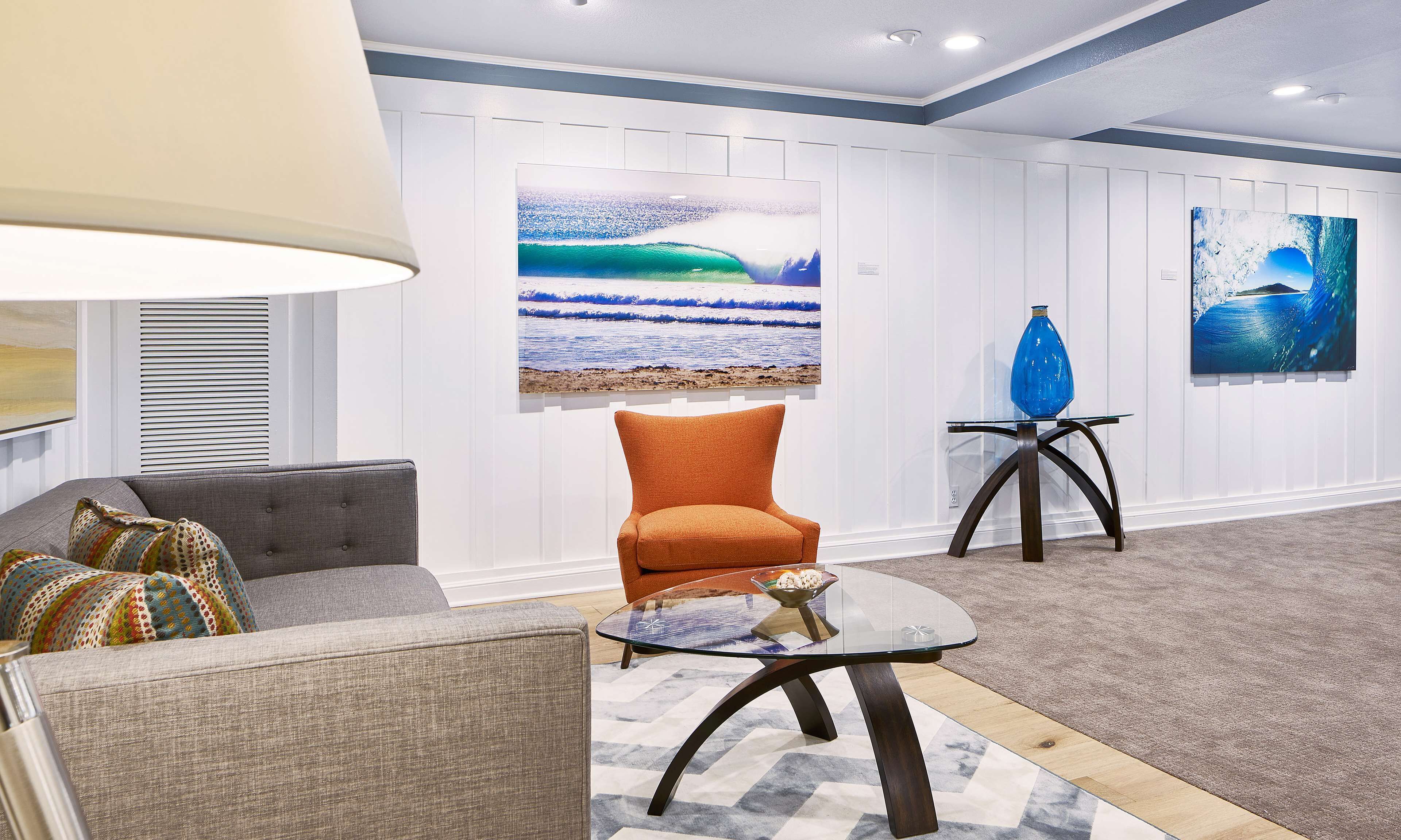A modern living room with a grey sofa, an orange armchair, a glass coffee table, and two paintings on the wall.