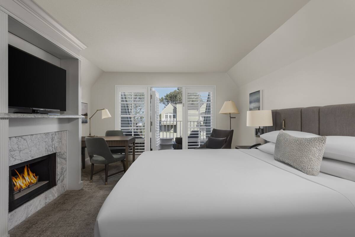 Habitación de hotel con una cama grande, televisión de pantalla plana, chimenea, zona de comedor con mesa y sillas, y balcón con vistas a edificios.