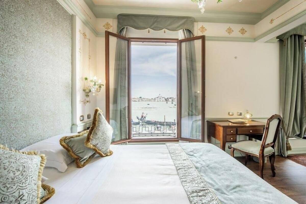 A hotel room with a large bed, a desk, and a view of a cityscape through a large window.