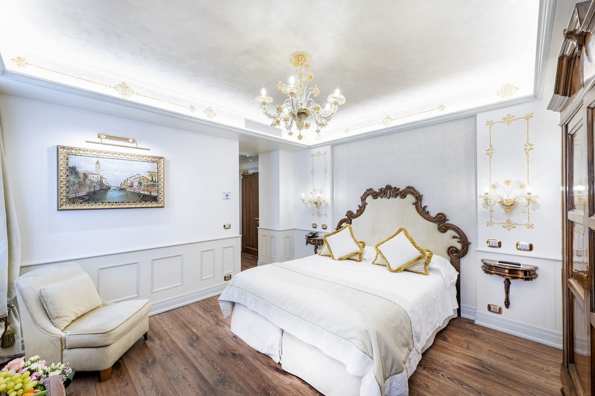 A bedroom with a large bed, a chandelier, a painting, and a wooden floor.