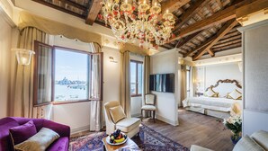 A spacious living room with a large chandelier, a sofa, a coffee table with fruits, and a view of a cityscape through large windows. - Hotel Monaco & Grand Canal (Venice)