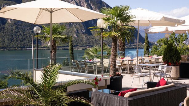 Terraza al aire libre con mesas y sillas, sombrillas y plantas en macetas, con vista a un lago y montañas.