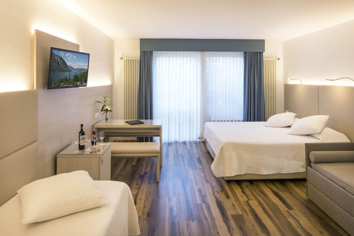 Ein Hotelzimmer mit zwei Betten, einem Schreibtisch, einem Stuhl, einem kleinen Tisch mit Flasche und Gläsern sowie einem Bild an der Wand.