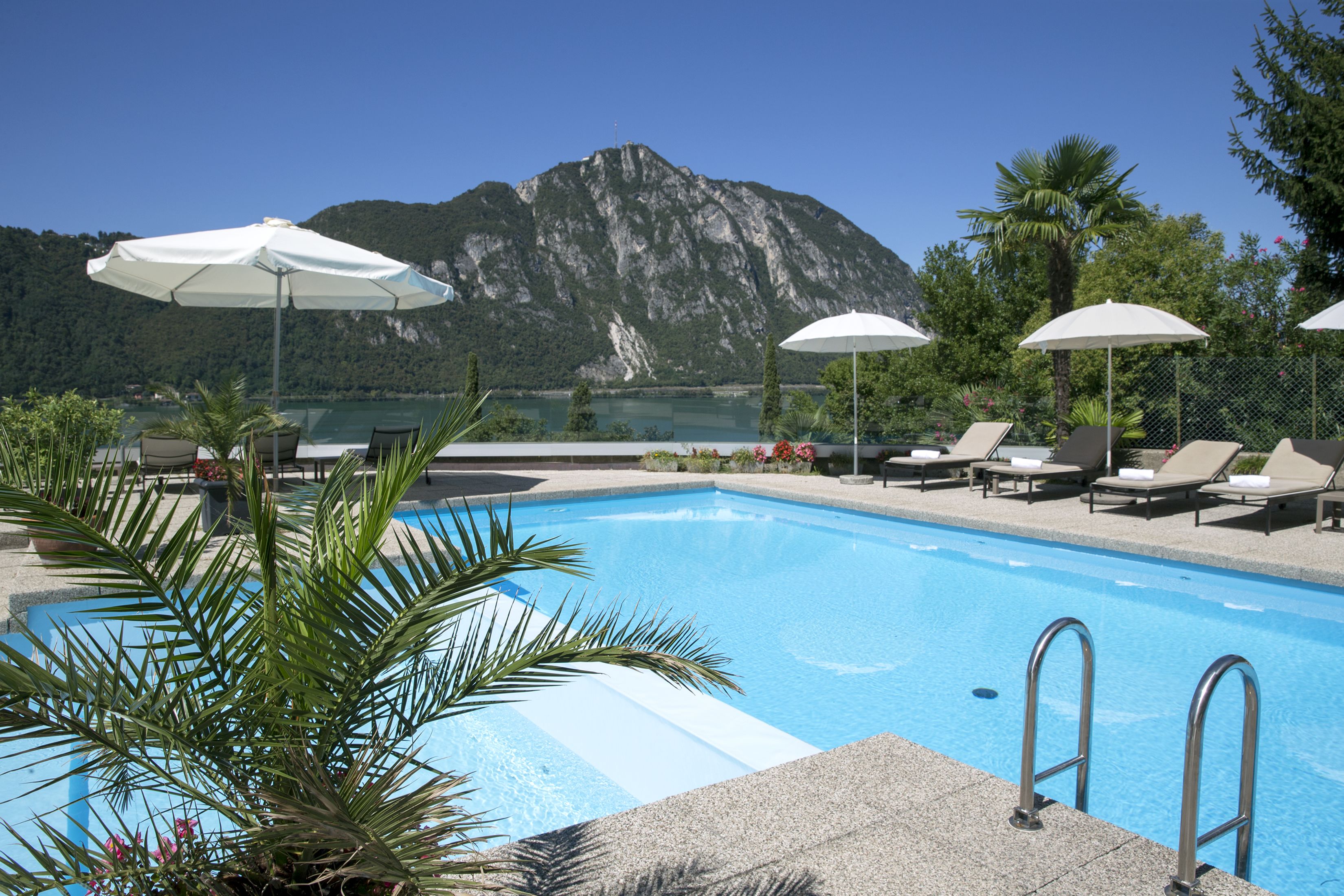 Ein Swimmingpool mit Liegestühlen und Sonnenschirmen, umgeben von Palmen und einem Berg im Hintergrund.