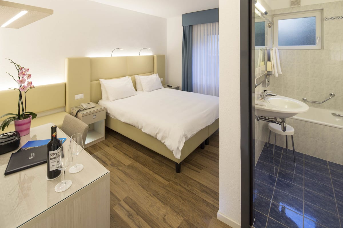Ein Hotelzimmer mit Bett, Nachttisch, Lampe, Zimmerpflanze und einem Badezimmer mit Waschbecken und Spiegel.